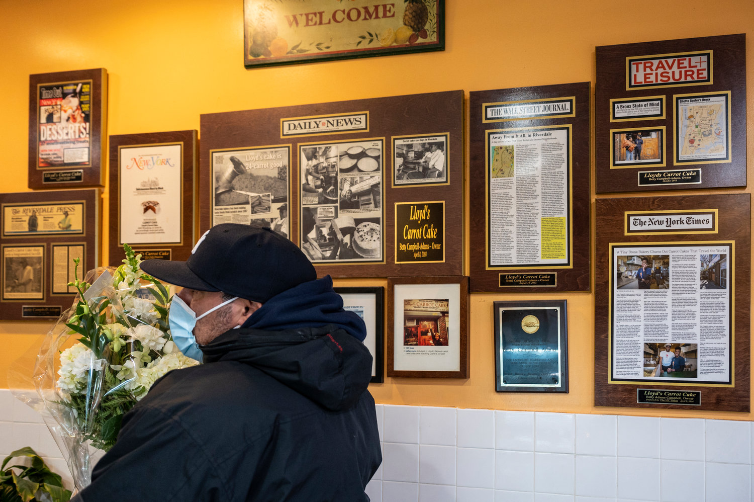 A man holds a bouquet of flowers intended for the grieving patrons of Lloyd’s Carrot Cake, standing in front of a wall celebrating the bakery’s media coverage from over the years. The bakery lost its co-founder, Betty Campbell-Adams, earlier this month.